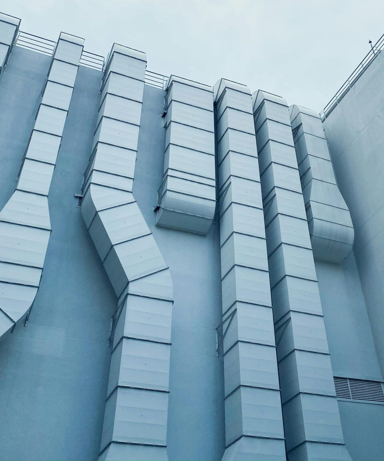 Low-angle view of industrial ductwork on a modern building exterior with sky and tree leaves.